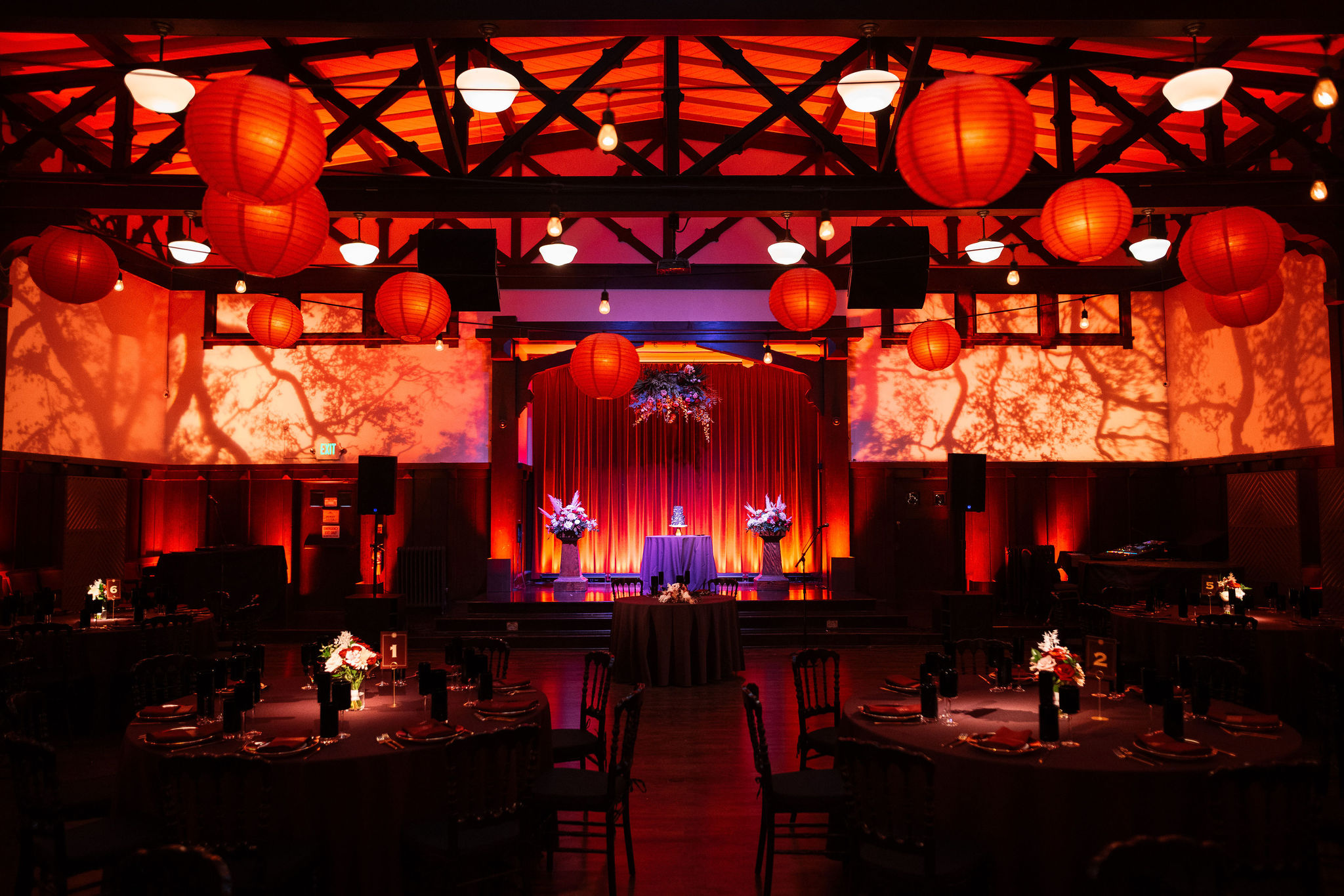 Wedding reception in the Swedish American Hall: round tables set for dinner beneath orange paper lanterns and warm red uplighting, tree-branch patterns projected across the walls, and a cake table flanked by floral arrangements on the curtained stage.