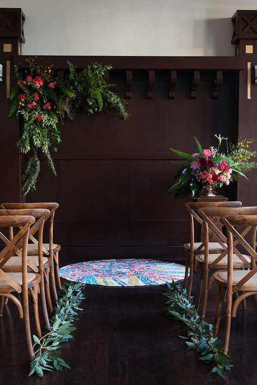 Decorated indoor wedding aisle with floral arrangements on dark wood backdrop and chairs on either side.