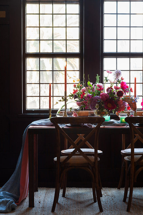 A wooden dining table with a floral centerpiece and candle holders with candles, in front of large grid-pane windows with sheer curtains.