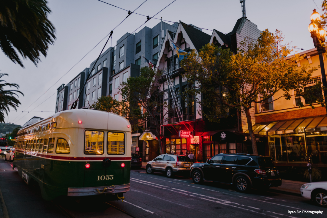 A street scene at dusk featuring a vintage trolleybus, parked cars, historic buildings with decorative facades, and a lit cafe sign.