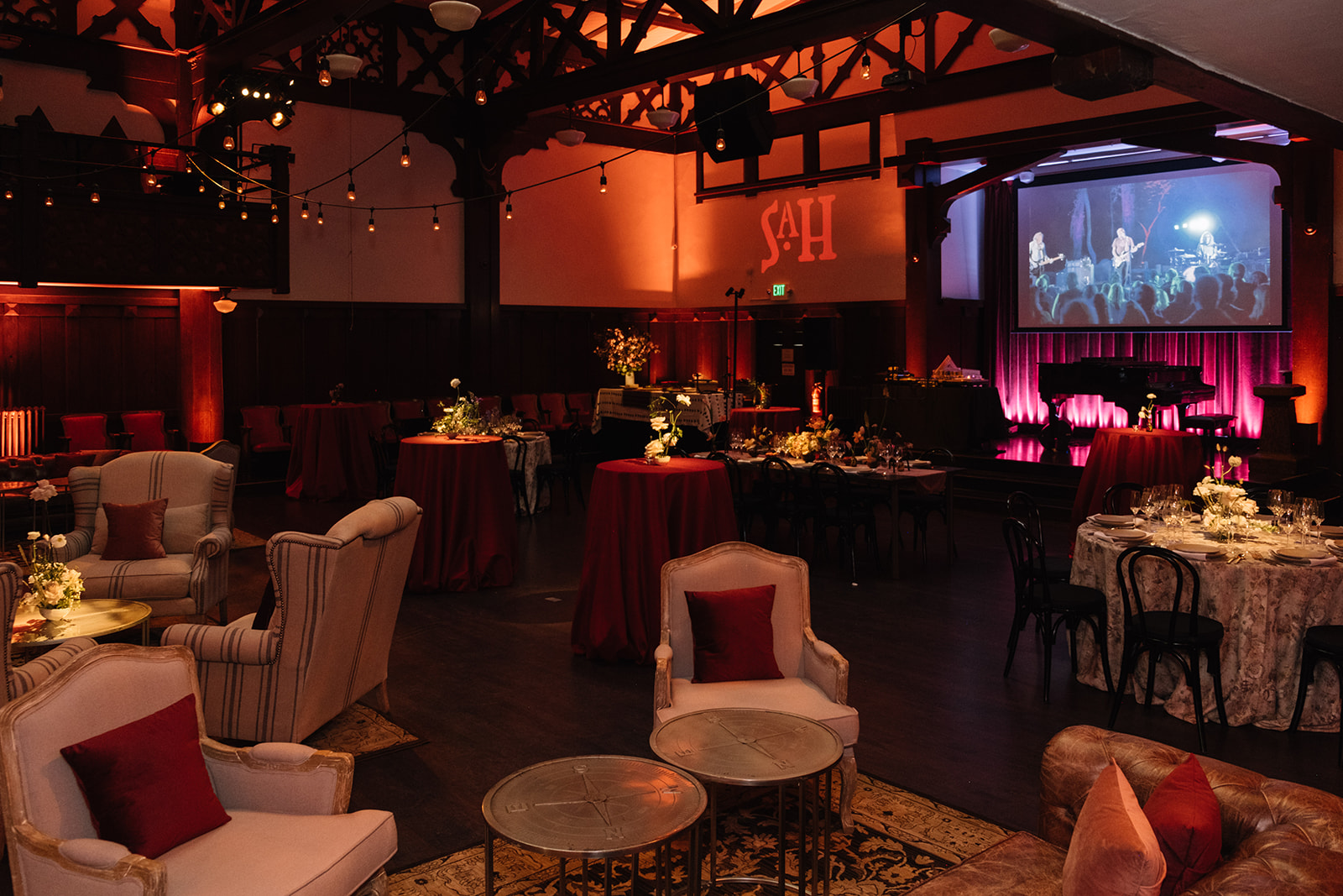 An elegant event space with a stage, a large screen displaying a band performance, cocktail tables with floral centerpieces, and cozy seating areas with armchairs and throw pillows, warm and ambient lighting.