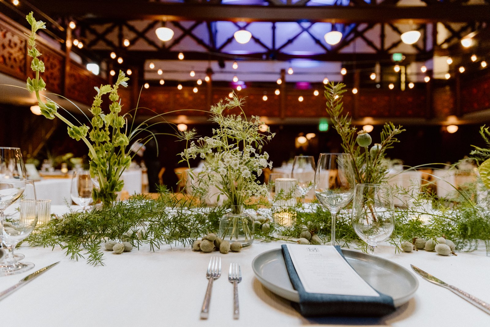 A decorated banquet table with glassware, plates, silverware, and floral centerpieces in a dimly lit event hall.