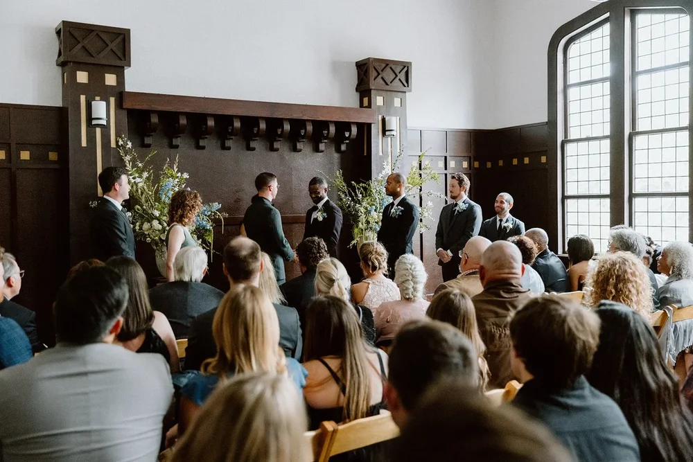 Guests seated for a wedding ceremony in a wood-paneled hall with tall windows.