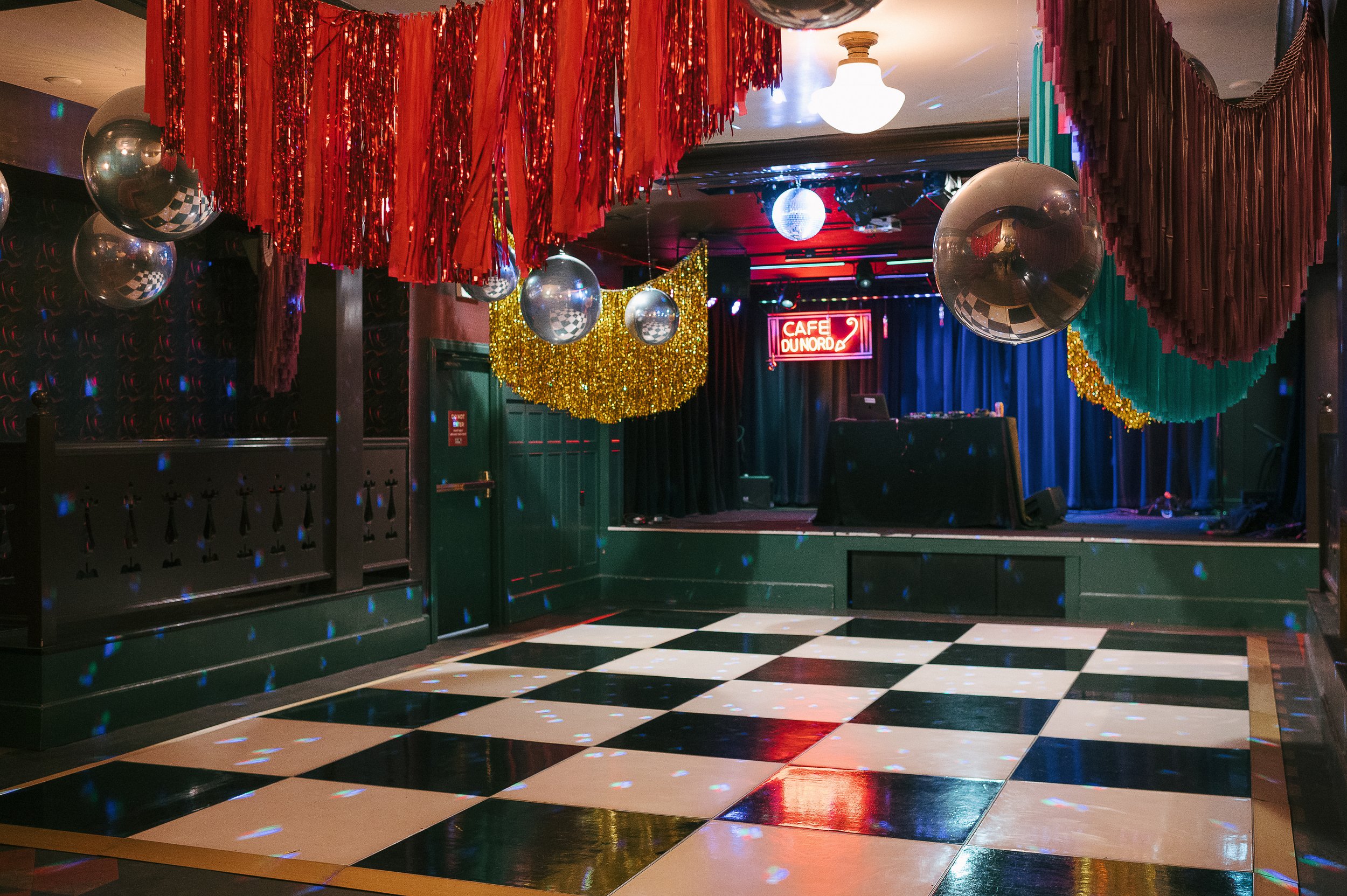 Empty dance floor with checkered black and white tiles, decorated for a party with red, gold, and green streamers, disco balls, and a stage with a DJ setup, neon lights, and a sign that reads 'Cafe Dunord'.