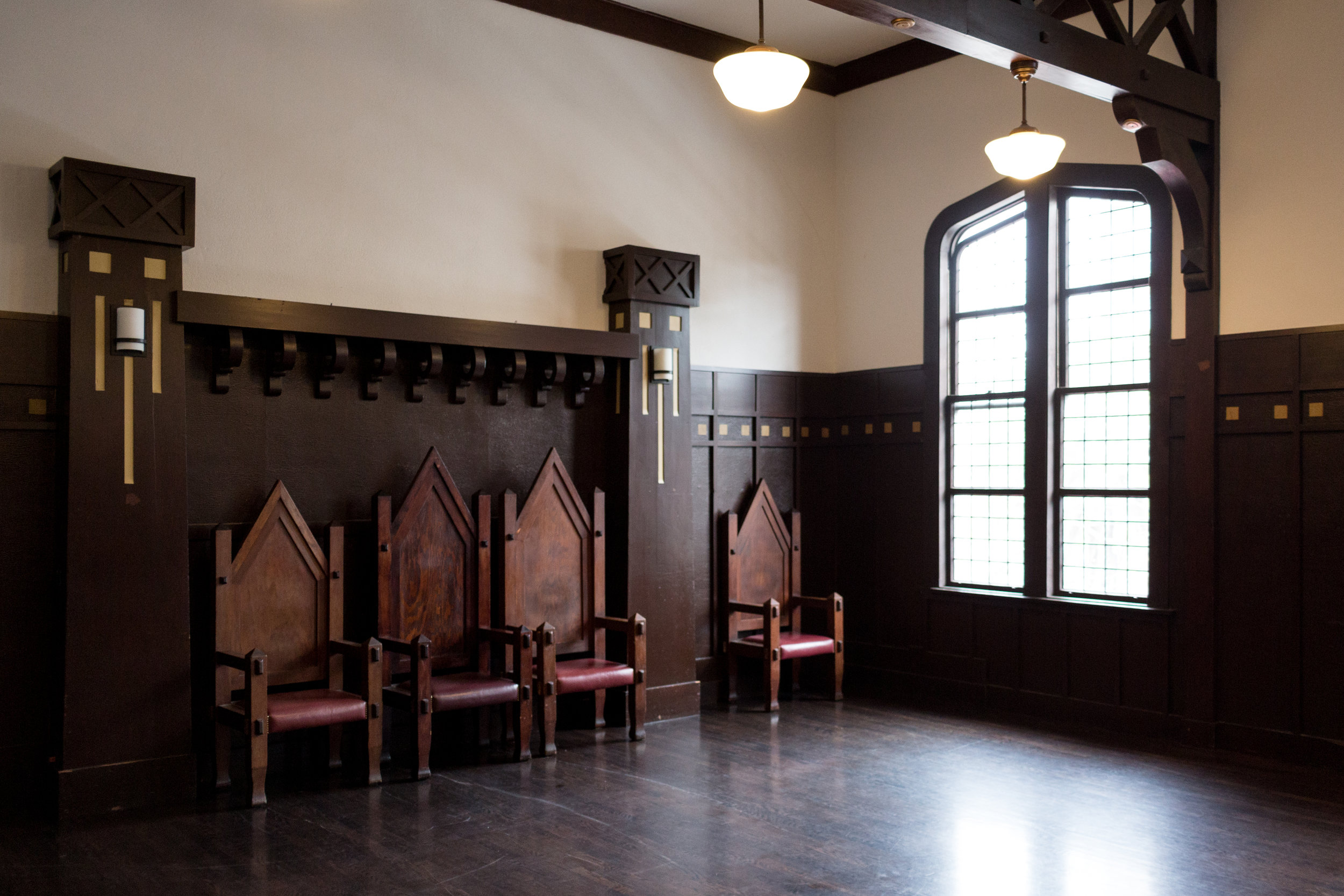 Empty room with dark wooden paneling, three chairs with tall spiked backs and red cushions, large arched window with grid pattern, and two ceiling lights.