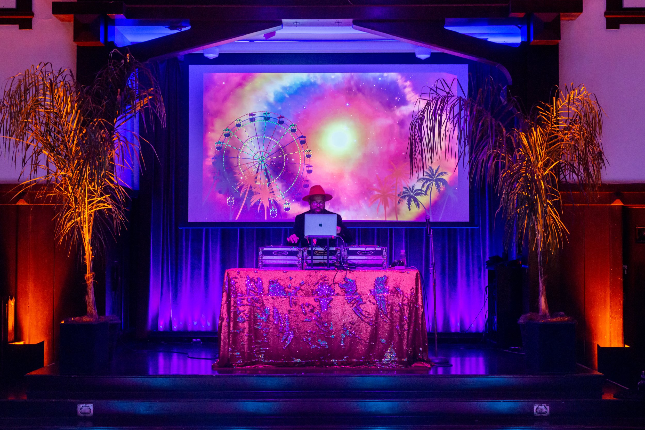 A DJ performing on stage with a colorful cosmic background on a large screen, featuring a Ferris wheel, palm trees, and stars. The DJ is wearing a red hat and glasses, with a laptop and equipment on a decorated table. Two large potted palm trees flank the stage, illuminated by purple and orange lighting.