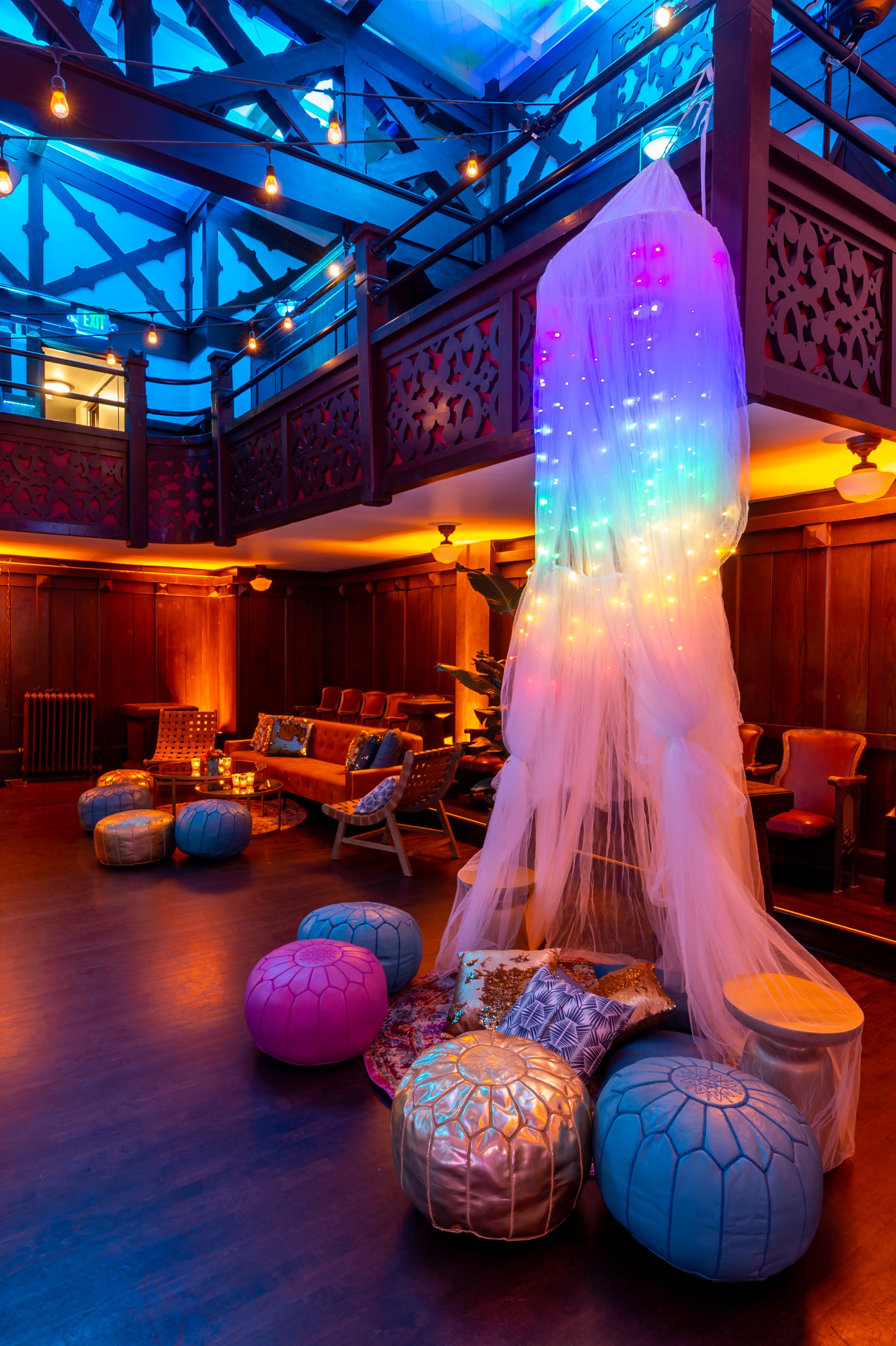 Interior of a vibrant lounge or bar with dim, warm lighting. Features a wooden wall, cozy seating, colorful poufs, and decorative pillows. A sheer, flowing fabric with rainbow-colored string lights hangs from the ceiling, creating a whimsical atmosphere. The upper level has a balcony with intricate wood railings and string lights hanging overhead.