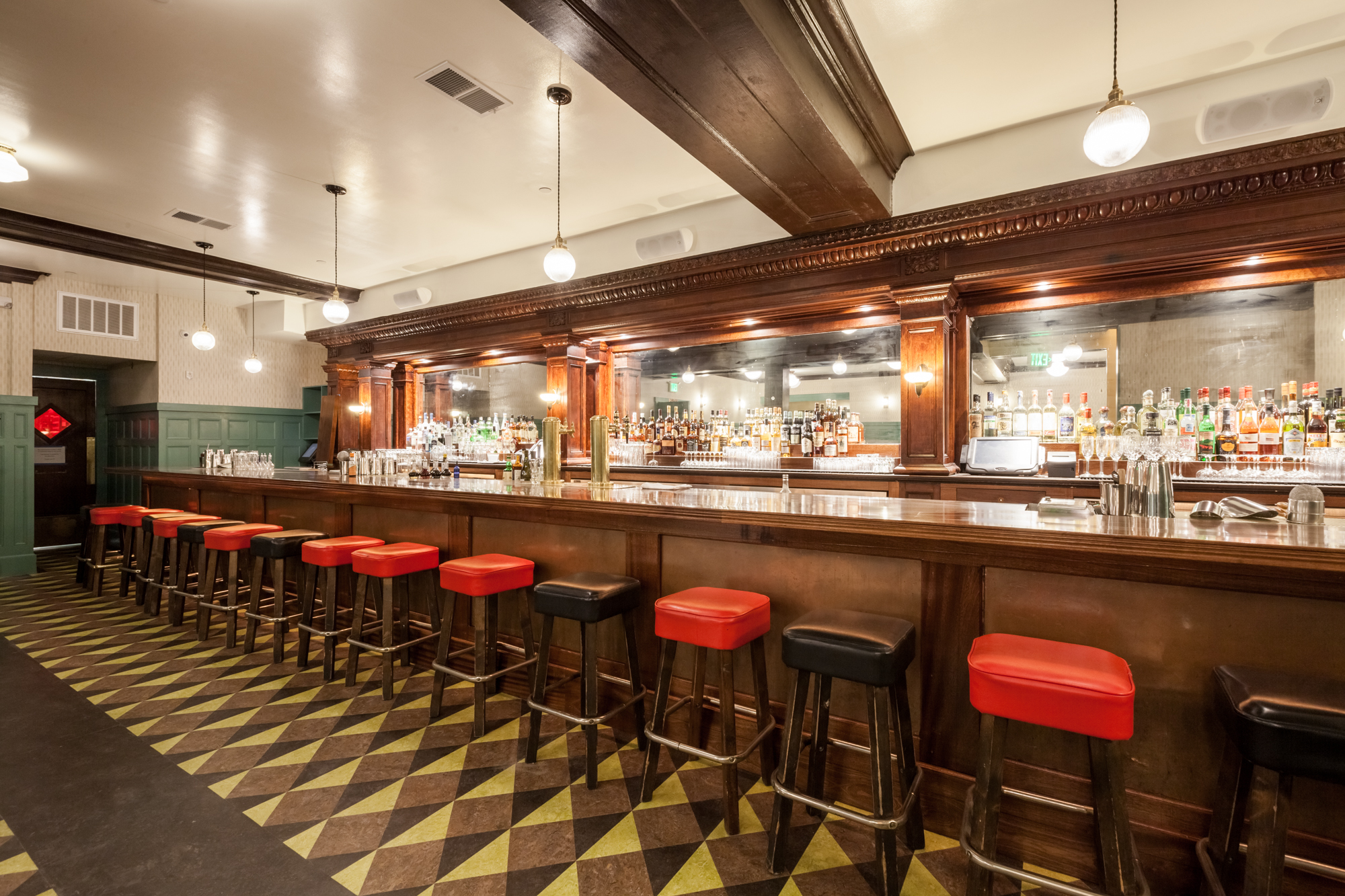 Empty bar with a wooden counter and red barstools in a dimly lit restaurant or bar interior. Shelves behind the counter are stocked with various bottles of alcohol and glasses.