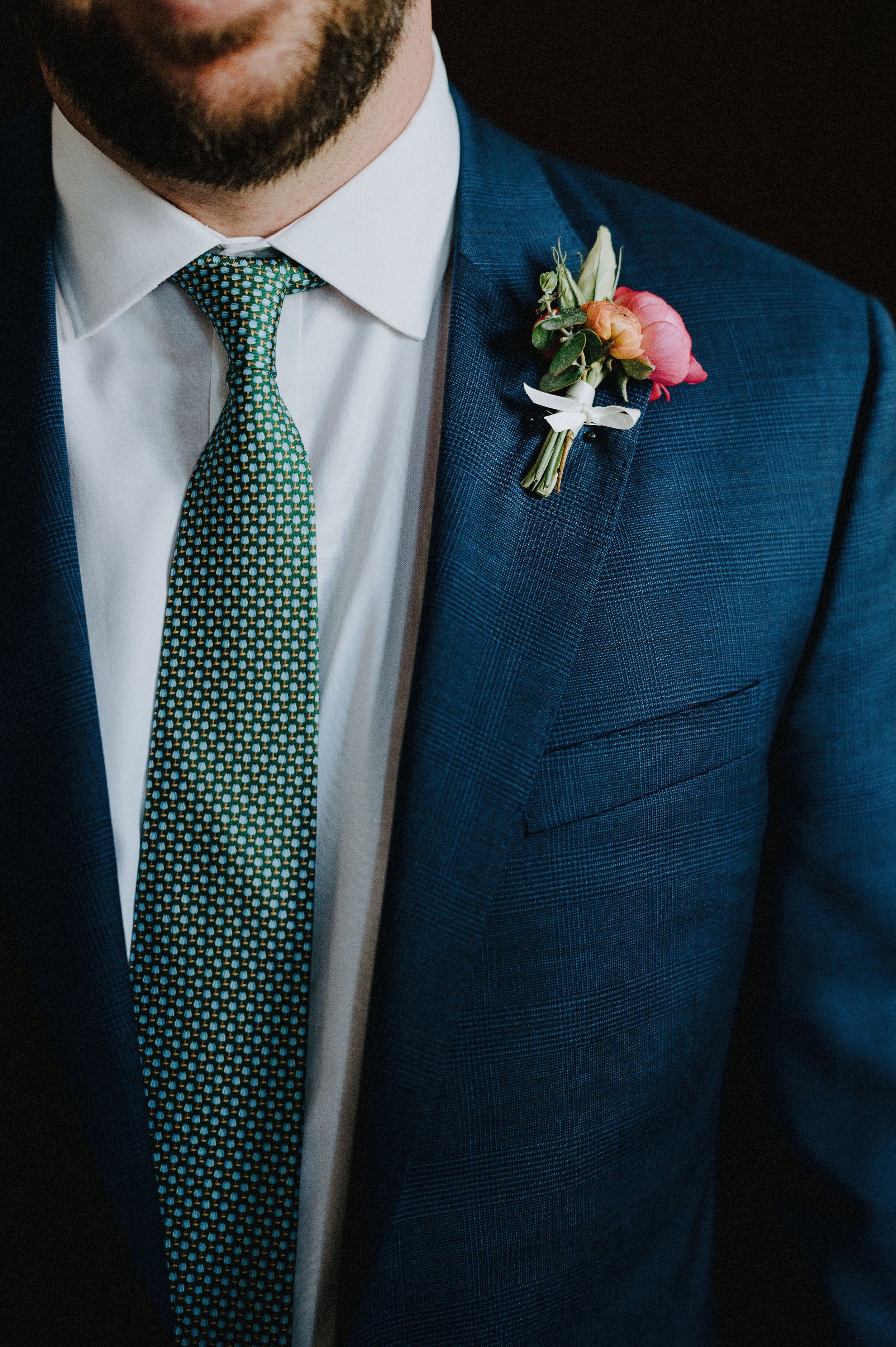 Close-up of a man wearing a dark blue checked suit, white shirt, patterned tie, and a boutonniere with pink and white flowers on his lapel.