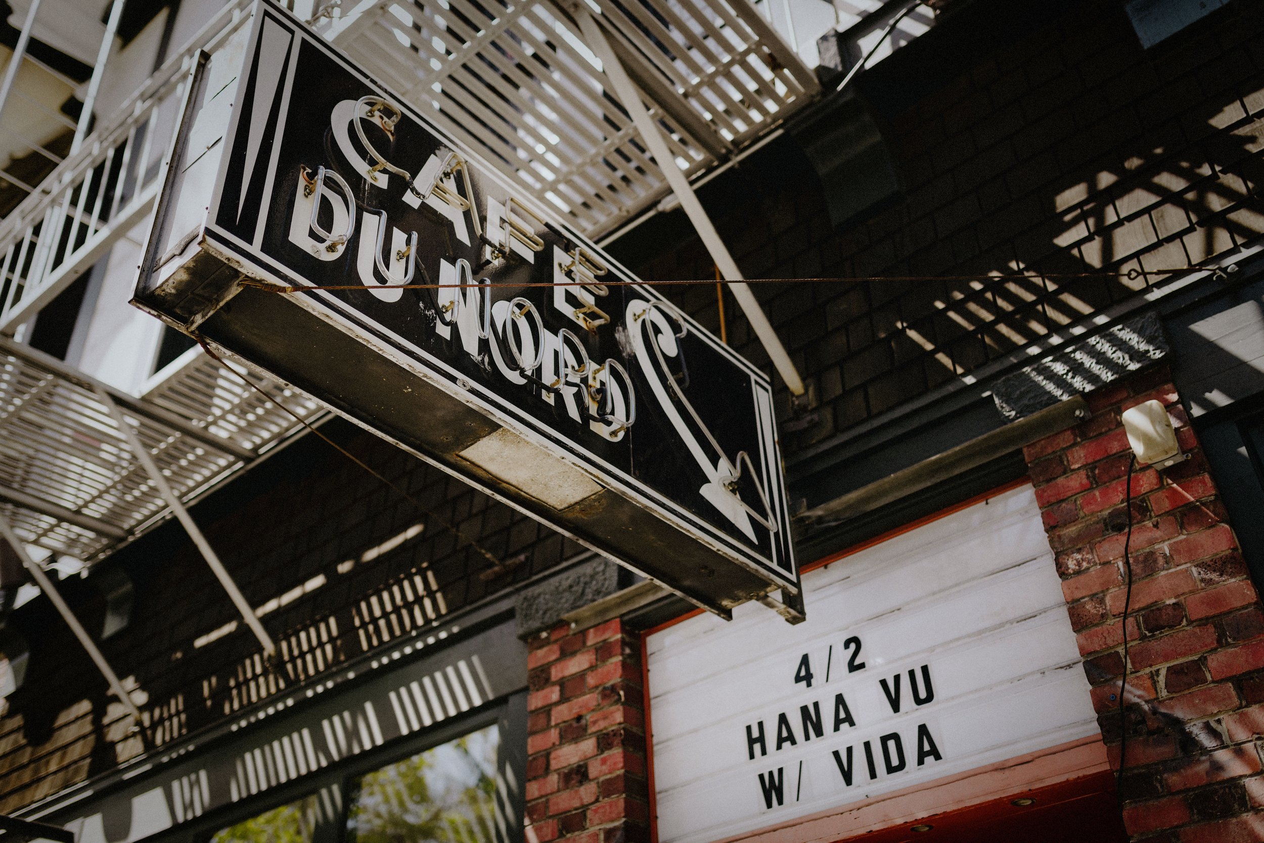 Neon sign that reads "CAFE DUBLIN" hanging above a brick building entrance with a white marquee sign displaying "4/2 HANA VU W/ VIDA".