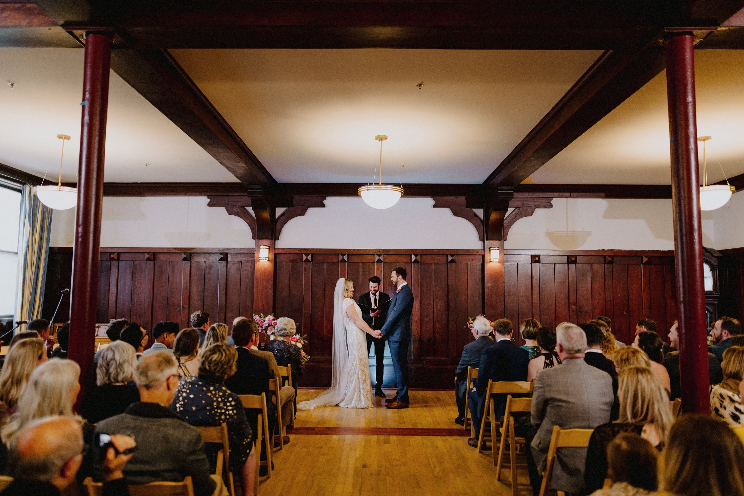 A wedding ceremony in a wood-paneled room with a bride and groom standing hand in hand and exchanging vows, officiated by an officiant, surrounded by seated guests.