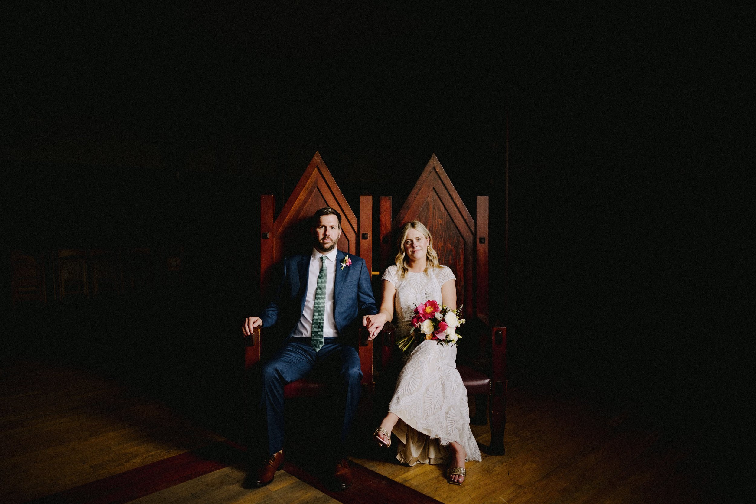 A bride and groom sitting on wooden chairs with high backs, holding hands. The bride is dressed in a white lace wedding dress and holding a bouquet of pink and white flowers. The groom is dressed in a dark blue suit with a white shirt and tie. The ba