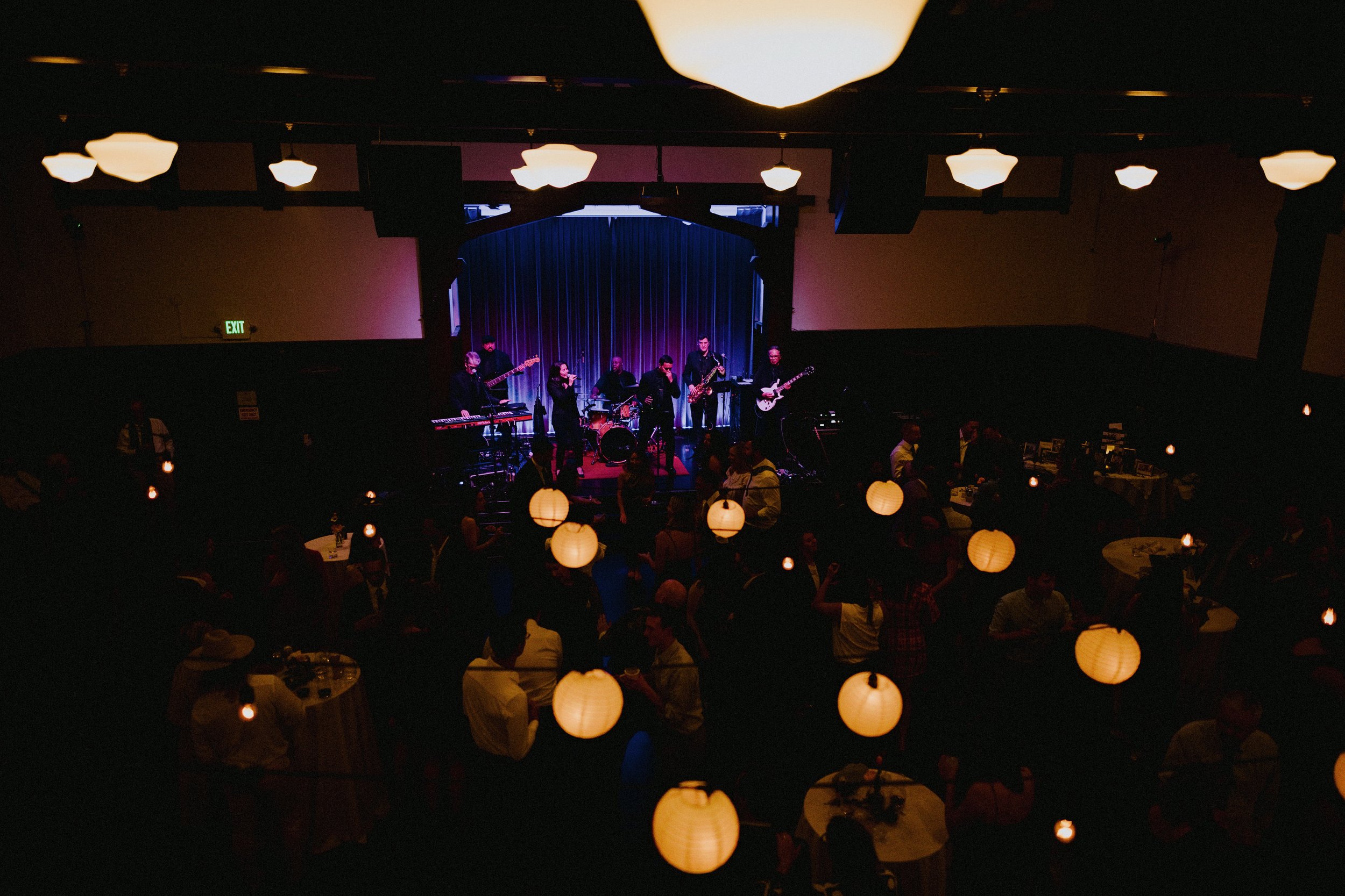 A band performing live on stage at an indoor concert with tables of people watching, illuminated by hanging paper lanterns.
