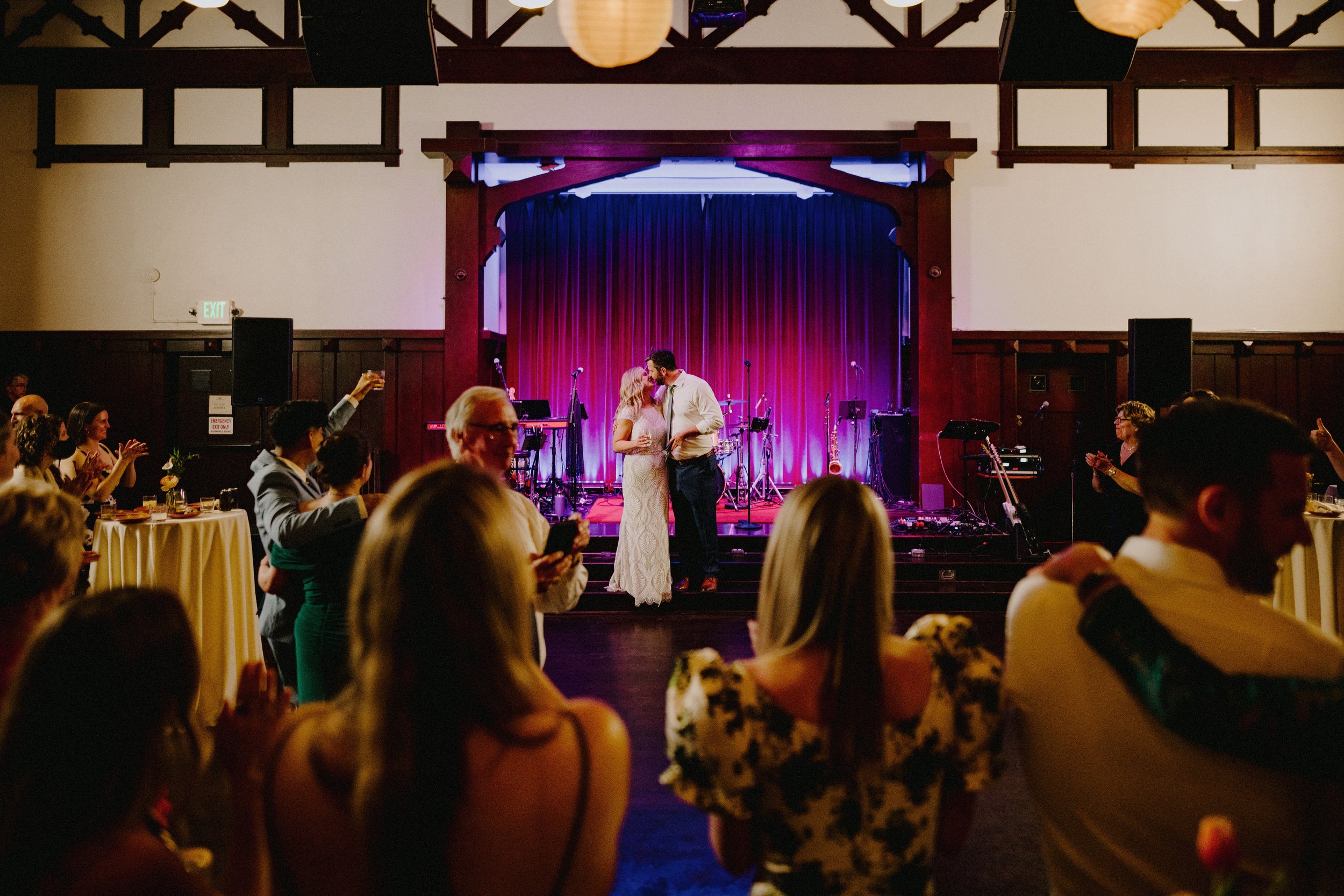 A couple shares a kiss on stage at a wedding reception, surrounded by guests, with a purple curtain and musical instruments in the background.