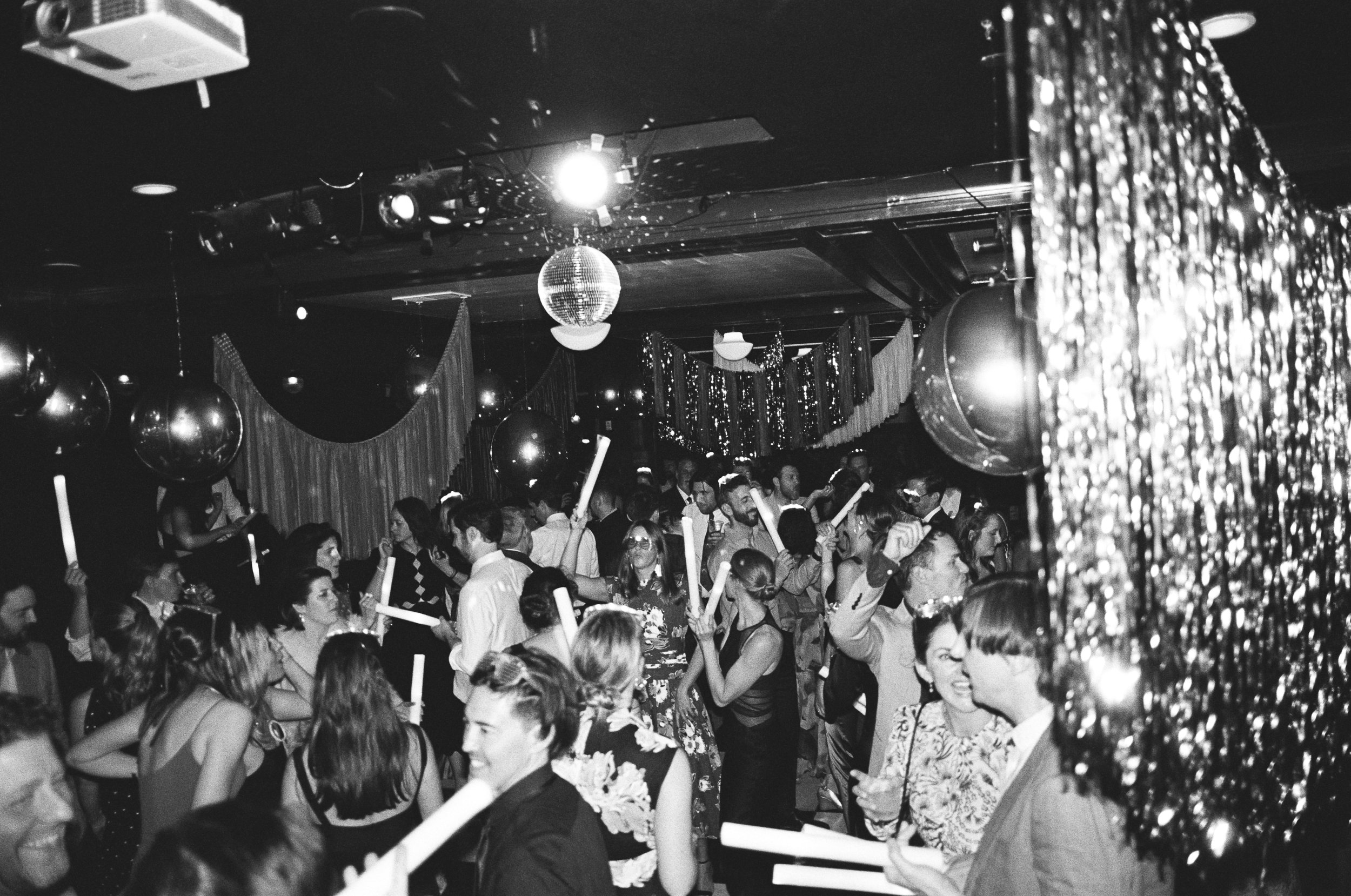 Crowd of people dancing and celebrating at a party with balloons and decorations, illuminated by a disco ball and stage lights in a festive setting.