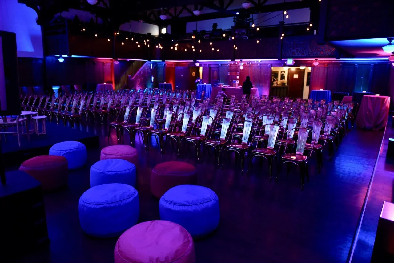 Empty event venue with rows of transparent chairs, colorful lighting, and round cushioned stools in the foreground.