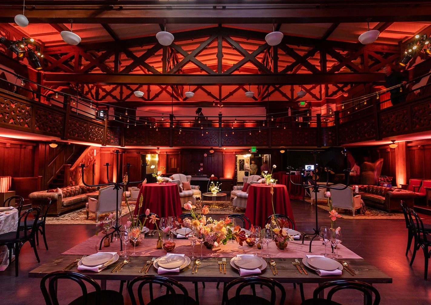 Elegant event space with red ambient lighting, floral centerpieces, and set dining tables in front of a sitting area with vintage sofas. The room has a high ceiling with exposed beams, a second-floor balcony with decorative wooden railing, and string lights hanging across the space.