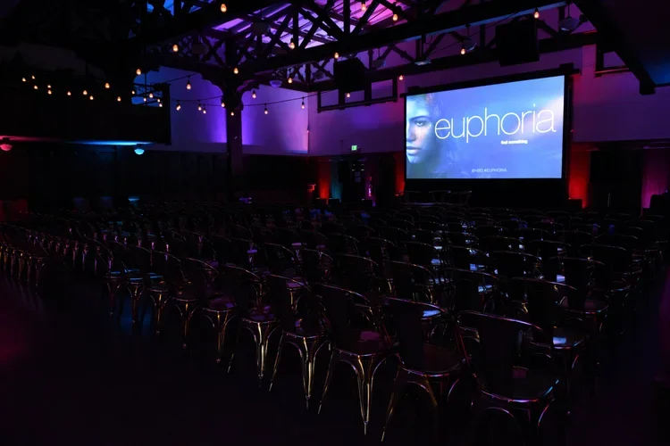 Empty theater or auditorium with rows of chairs, a large projection screen displaying the word 'euphoria' and an image of a woman's face, and stage lighting creating a purple and red glow.