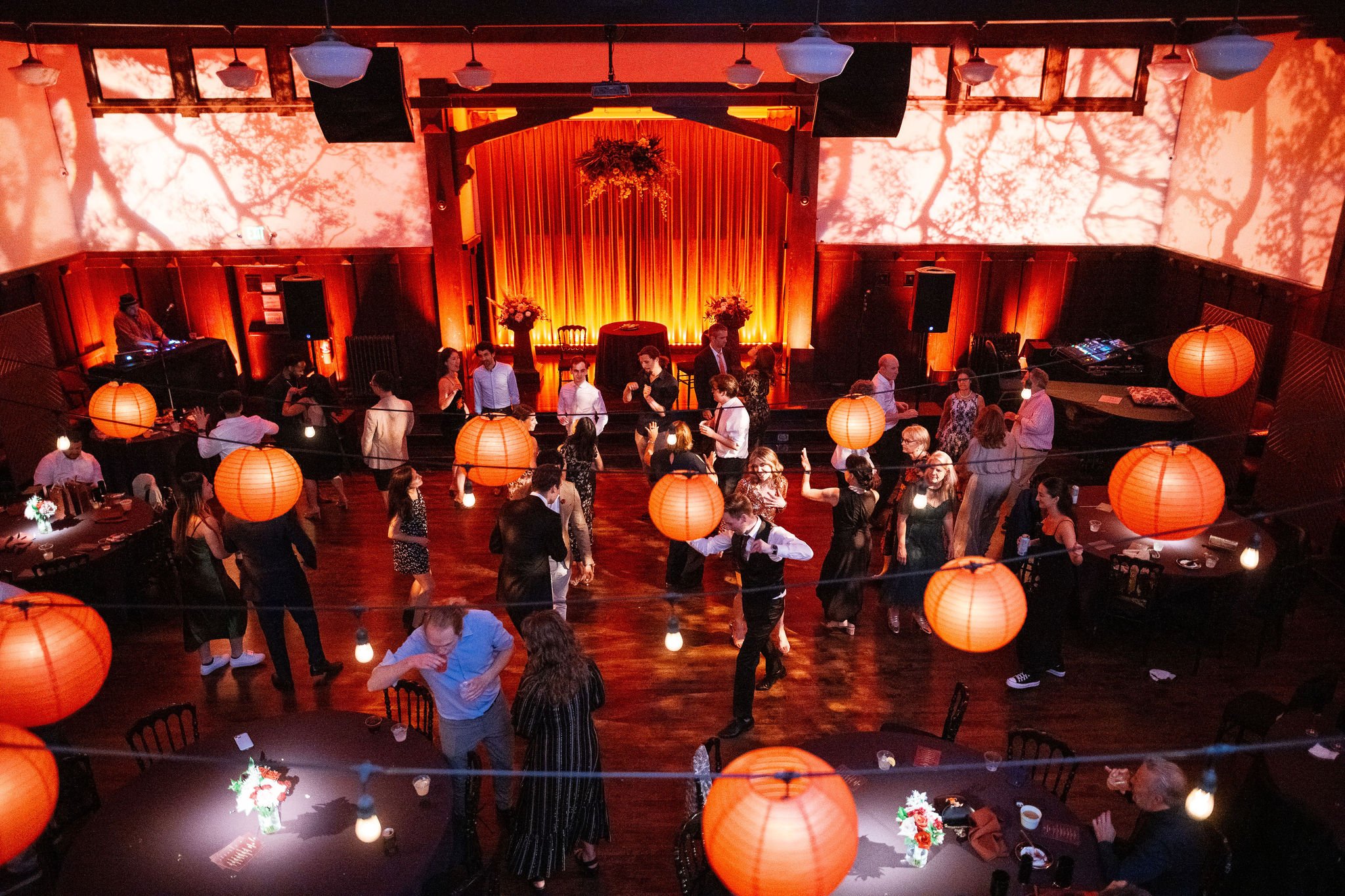 Indoor event space with warm orange lighting, hanging paper lanterns, and people socializing around tables with floral centerpieces.