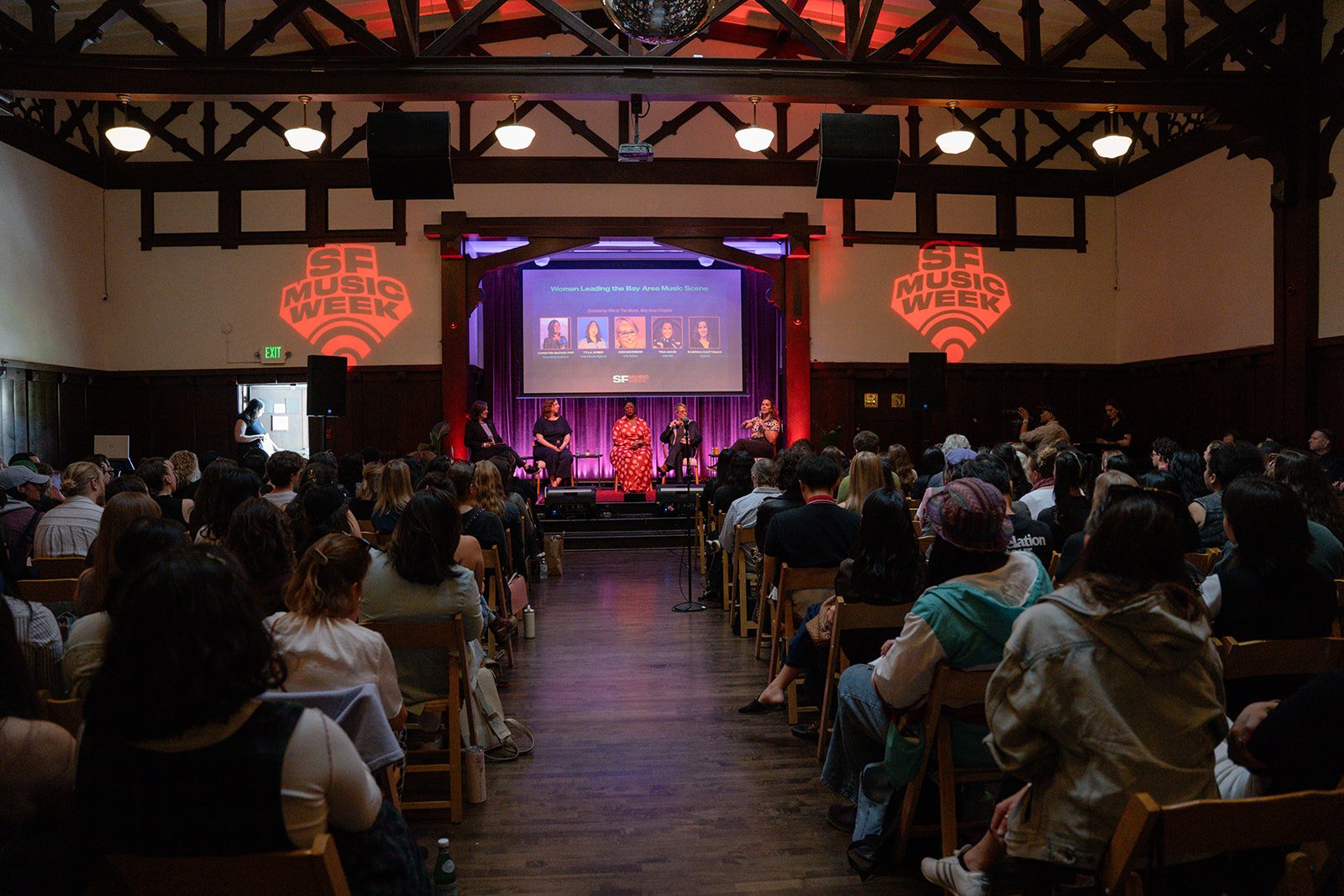 A panel discussion at a music event with five women seated on stage, a large audience in front, and the words 'SF Music Week' projected on the walls.