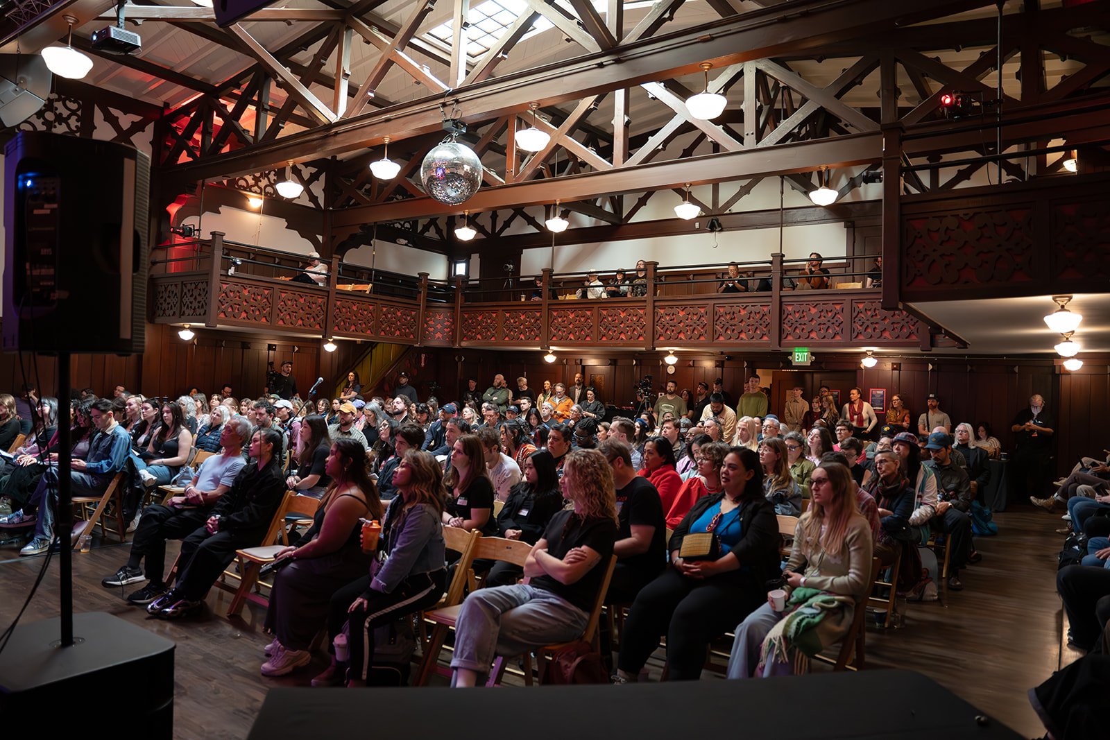 An audience seated in a wooden auditorium watching a presentation or performance, with people on the upper balcony level.
