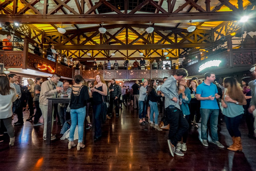 People socializing and dancing inside a large, wooden-beamed venue with a second-floor balcony and warm lighting.