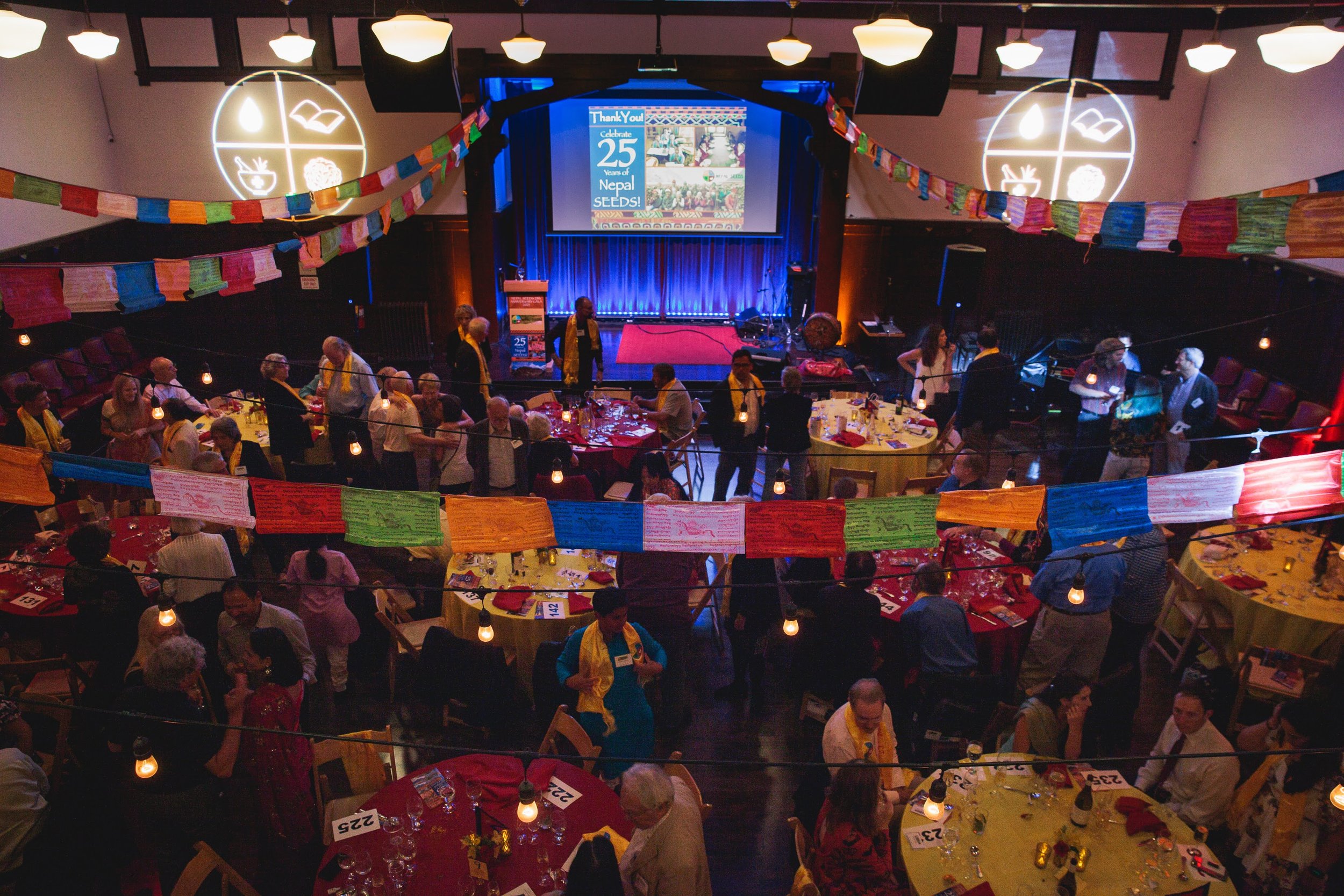 People gathered at tables with yellow and red tablecloths in a decorated event space, celebrating Nepal's 25 years of seed development, with colorful prayer flags, a stage with a projector screen, and illuminated symbols on the wall.