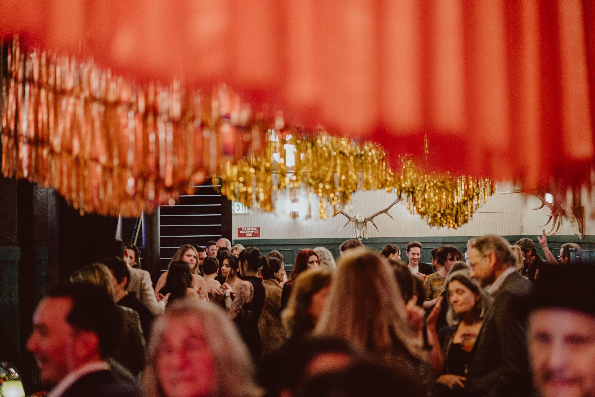 A crowd of people gathered indoors beneath hanging gold and red decorations, with a deer skull mounted on the wall in the background.