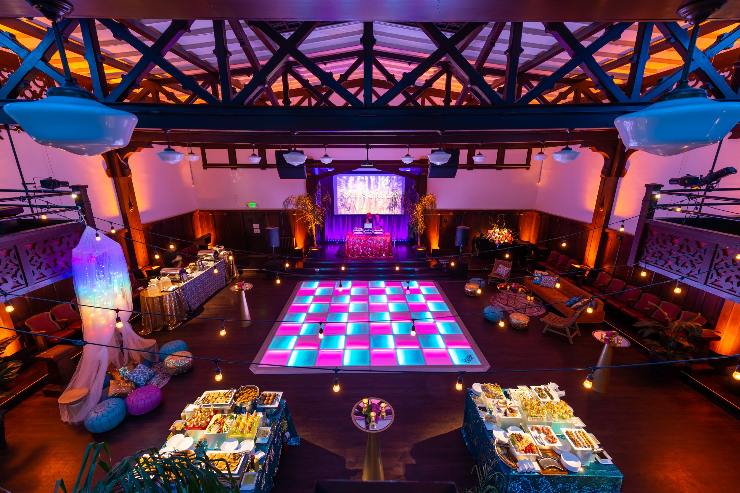 Interior of a decorated event hall with a colorful dance floor, buffet tables, a DJ setup, and seating areas, illuminated by vibrant lighting and string lights.