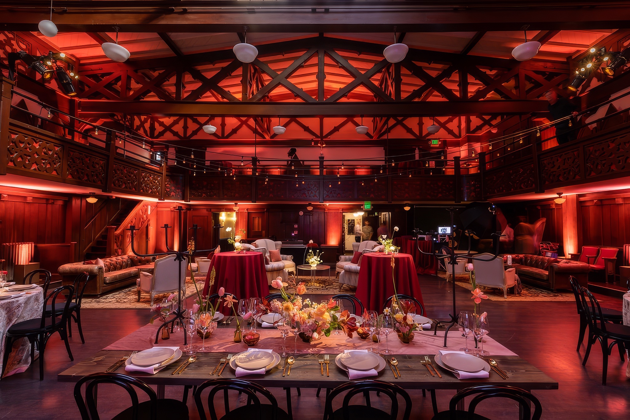 The Swedish American Hall lit in deep red, with a long banquet table set beneath the timbered ceiling, lounge seating and cocktail tables on the floor, and the carved wooden balcony above.