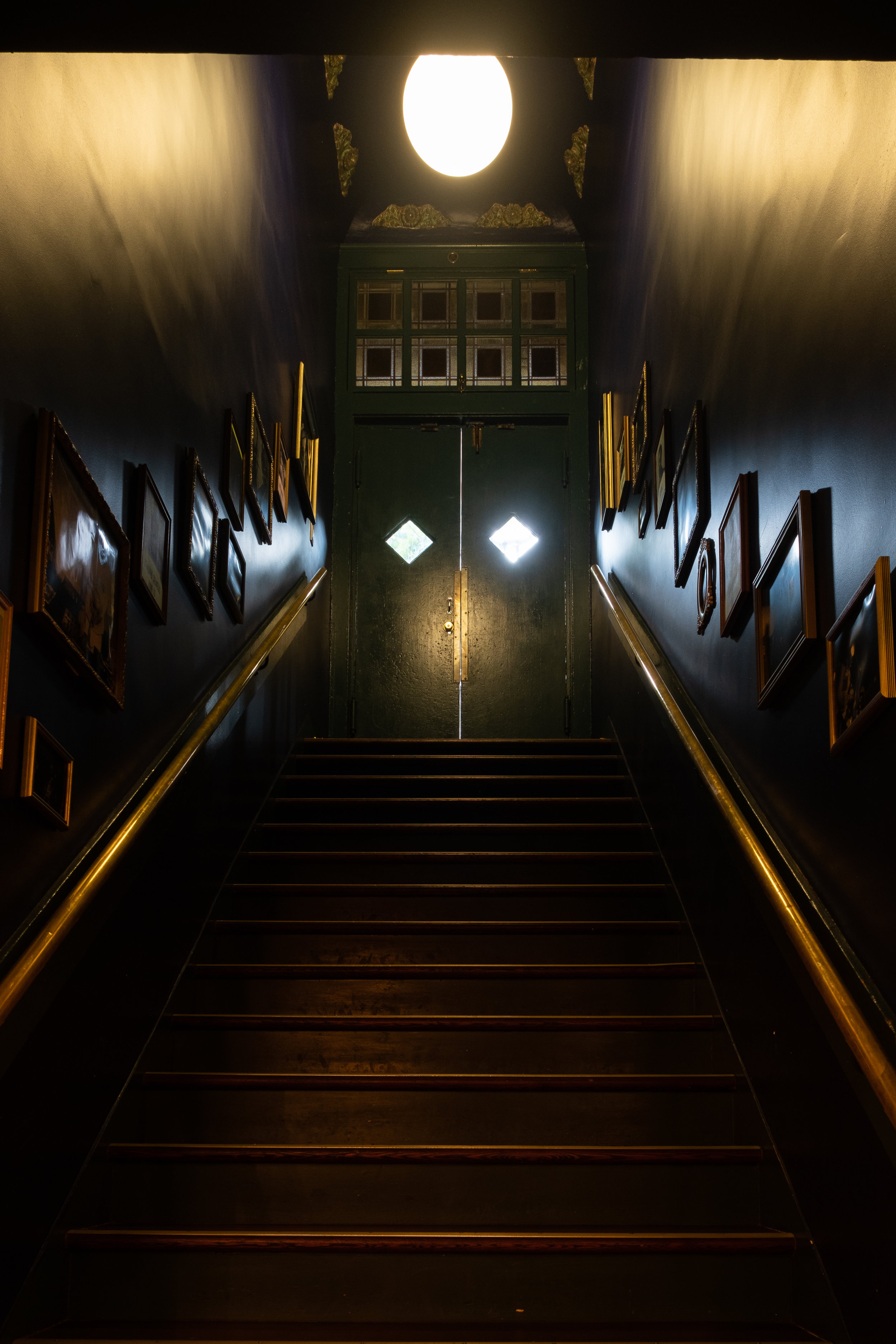 A staircase leading up to a green door with diamond-shaped windows, framed by dark walls decorated with framed pictures and illuminated by a ceiling light.