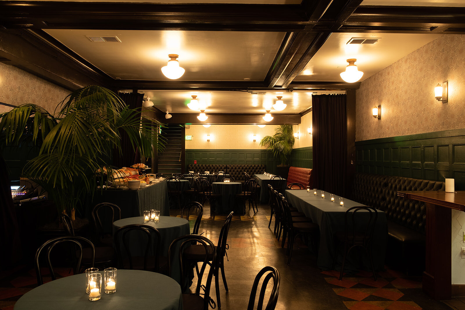 Elegant dimly lit restaurant with round tables, candles, and black chairs, decorated with potted plants and vintage wallpaper.