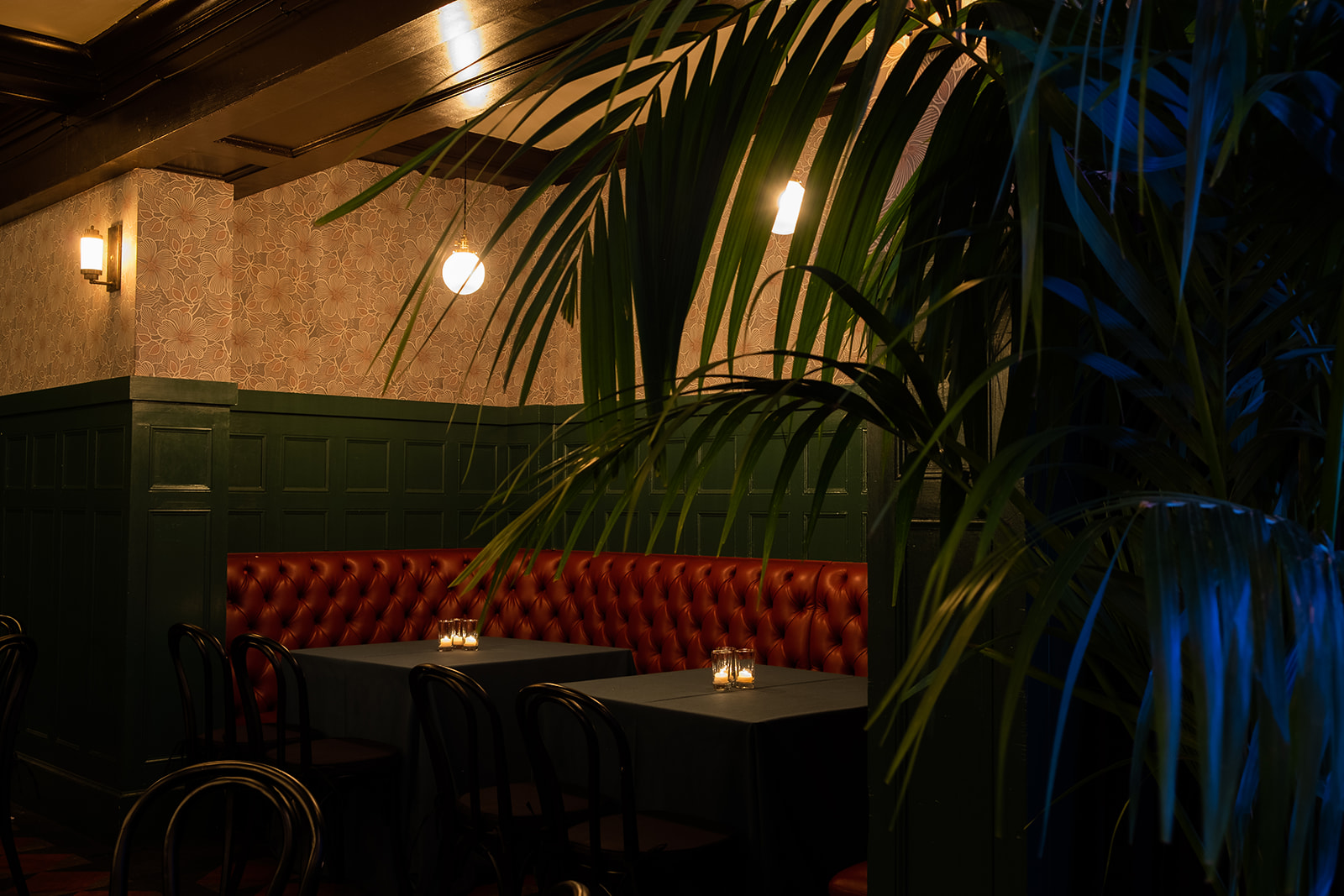 Dimly lit restaurant corner with dark tables, black chairs, red leather booth seating, green paneled walls, floral wallpaper, and hanging lights, partially obscured by green foliage.