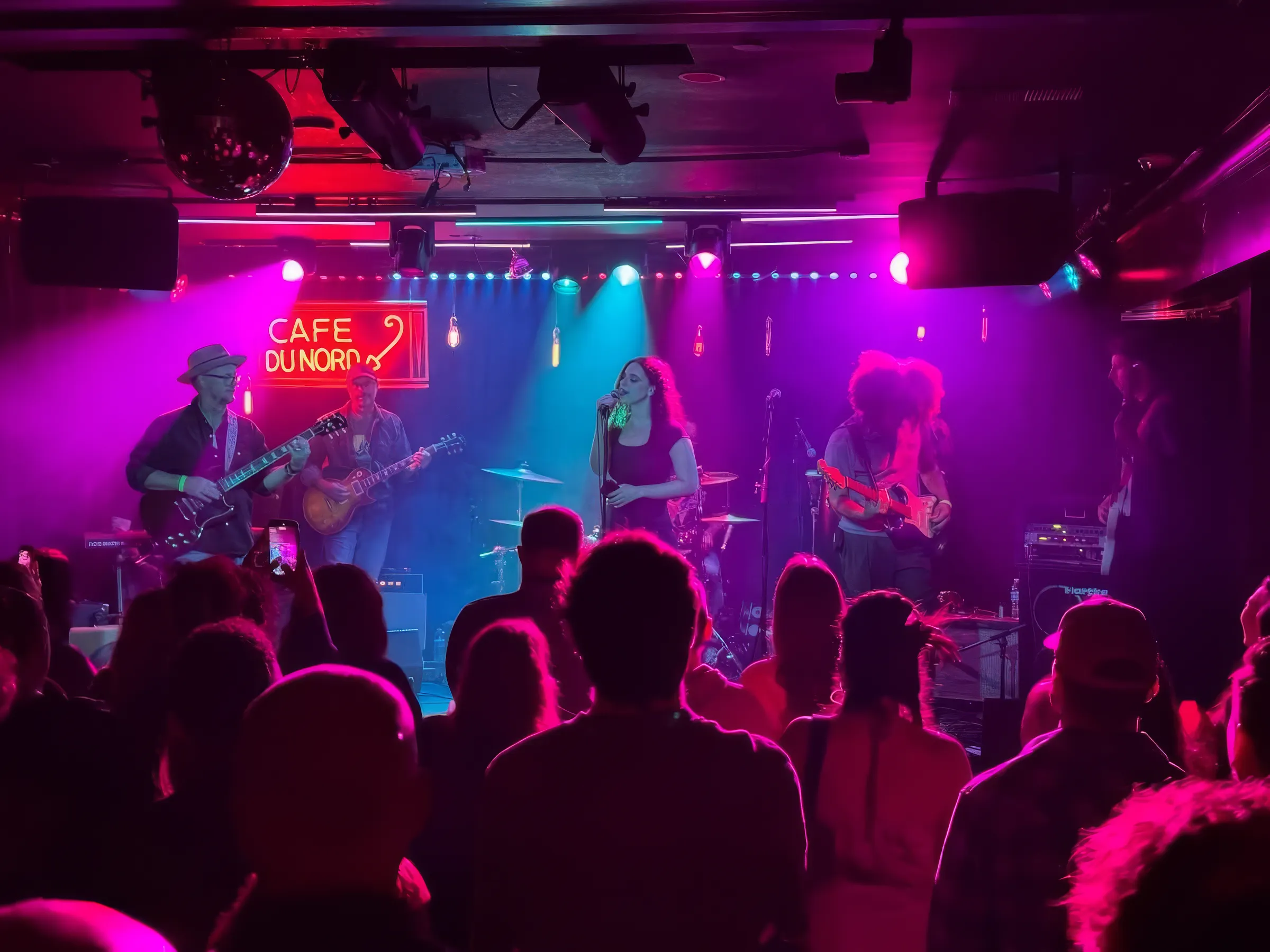 A band playing the Cafe du Nord stage beneath the venue's red neon sign, lit in magenta and cyan, with a full crowd watching from the floor.