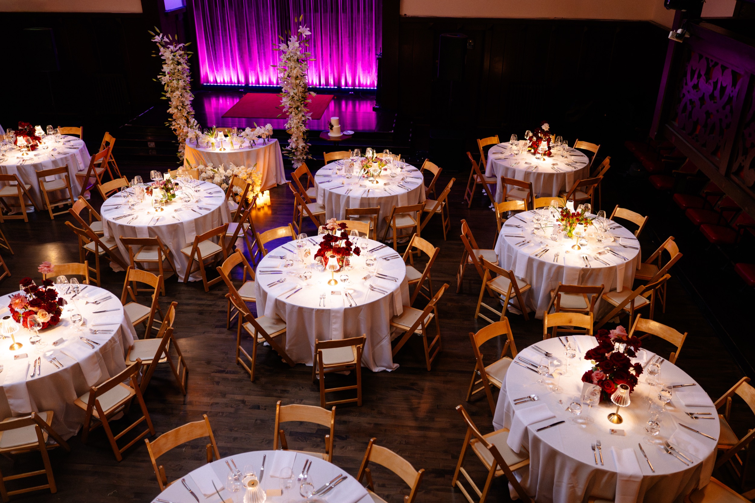 Round banquet tables in white linen with red and blush floral centerpieces, viewed from the Swedish American Hall balcony, with a magenta-lit stage and cake table beyond.